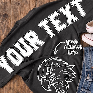 May include: A black t-shirt featuring the white outlined text YOUR TEXT in a bold varsity font, a white line drawing of an eagle head, and the white cursive text your mascot here with a white arrow pointing toward the eagle graphic.