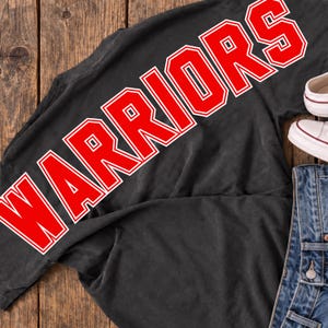 May include: A black t-shirt featuring the word WARRIORS printed in large, bold red letters with a white outline. The shirt is positioned next to a pair of white canvas sneakers and blue denim jeans, showcasing a casual, everyday ensemble.