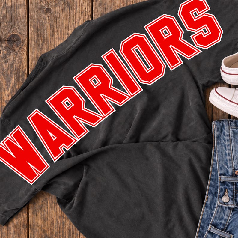 May include: A black t-shirt featuring the word WARRIORS printed in large, bold red letters with a white outline. The shirt is positioned next to a pair of white canvas sneakers and blue denim jeans, showcasing a casual, everyday ensemble.