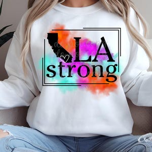 以下が含まれることがあります： 白いスウェットシャツに、カリフォルニア州の黒いアウトラインと、「LA strong」という黒いテキストが描かれており、「A」の中に白いハートがあります。テキストは、ピンク、オレンジ、ブルー、ターコイズの色の水彩画のデザインで囲まれています。