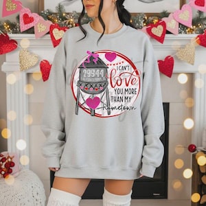 Puede incluir: Sudadera gris claro con un gráfico de una torre de agua, corazones y el texto "I can't love you more than my hometown". La torre de agua tiene el texto "29544 Galivants Ferry, SC."