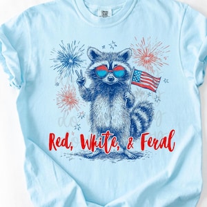 Peut inclure: T-shirt bleu clair avec un raton laveur portant des lunettes de soleil et tenant un drapeau am&eacute;ricain. Le raton laveur fait le signe de la paix. Le texte "Red, White, & Feral" est &eacute;crit en rouge en dessous.