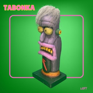 May include: A purple and green hand-painted wooden figurine with a white hairpiece. The figurine has a large nose, yellow eyes, and a toothy grin. The figurine is standing on a black base and has the word "TABONKA" written in yellow letters on the top.