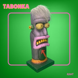 May include: A purple and gray ceramic sculpture of a face with a white hairpiece. The face has a large nose, yellow eyes, and a wide mouth with gold teeth. The sculpture is on a black base and has the word "TABONKA" written in yellow letters on a pink background.