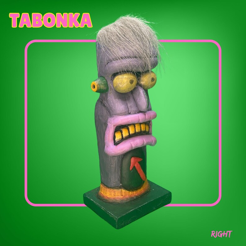 May include: A purple and gray ceramic sculpture of a face with a white hairpiece. The face has a large nose, yellow eyes, and a wide mouth with gold teeth. The sculpture is on a black base and has the word "TABONKA" written in yellow letters on a pink background.