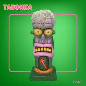 May include: A purple and green wooden tiki statue with a white hairpiece, yellow eyes, and a pink mouth with yellow teeth. The statue has a red arrow on its chest and the word "TABONKA" is written in yellow letters at the top of the image.