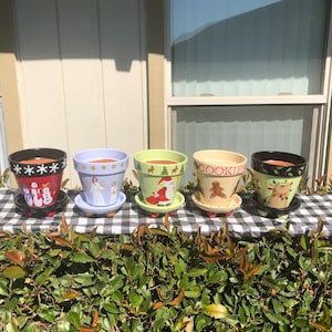 Puede incluir: Cinco macetas de terracota pintadas a mano con motivos navideños. Las macetas están decoradas con un muñeco de nieve, un ángel, Papá Noel, un hombre de jengibre y un reno. Cada maceta tiene un platillo.