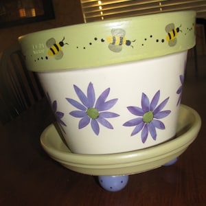 May include: A hand-painted ceramic flower pot with a green rim and a white base. The pot is decorated with purple flowers and yellow and black bees. The pot sits on a green saucer with three blue feet.