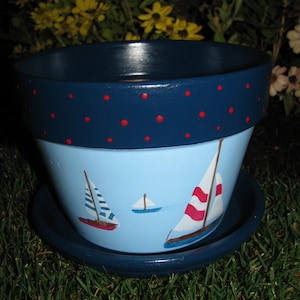 May include: A blue and white ceramic planter with a red polka dot trim. The planter is decorated with a blue sky background and three white sailboats with red and blue accents.