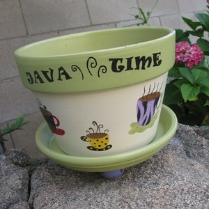 May include: A green and white ceramic planter with the words "Java Time" painted on the side. The planter has a saucer and features a whimsical design of coffee cups and swirls.