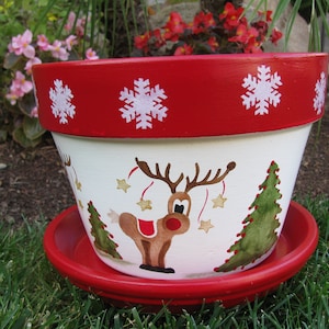 May include: A white ceramic planter with a red rim and a painted reindeer design. The planter has a red saucer and is decorated with white snowflakes.