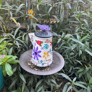 May include: A white, floral-painted bird feeder with a black polka-dotted lid and a purple butterfly handle. The feeder is filled with birdseed and sits on a gray, circular tray, surrounded by green foliage.