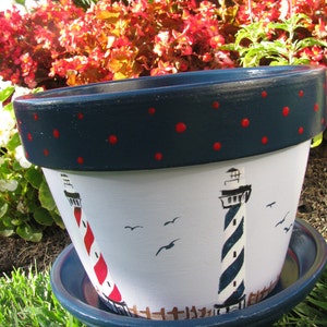 May include: A blue and white ceramic planter with a lighthouse design. The planter has a dark blue rim with red polka dots and a white base with two lighthouses painted on it. The planter is sitting on a bed of green grass.
