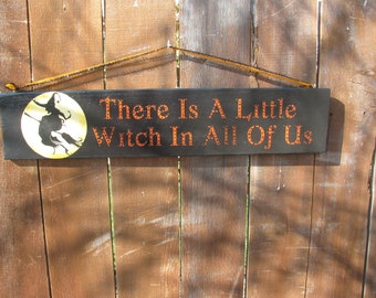 May include: Black wooden sign with a witch silhouette flying on a broom in front of a yellow moon. The sign reads "There is a little witch in all of us".