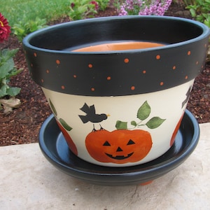 May include: A black and white painted ceramic planter with a black rim and orange polka dots. The planter features a painted pumpkin with a jack-o'-lantern face and a black bird perched on a branch.