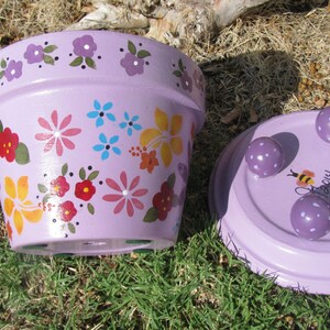May include: Purple flower pot with a painted floral design and a matching saucer with three feet. The saucer has a bee and the text "Happy" painted on it.