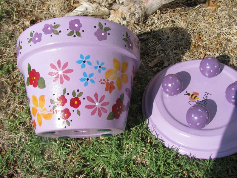 May include: Purple flower pot with a painted floral design and a matching saucer with three feet. The saucer has a bee and the text "Happy" painted on it.