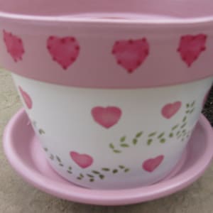May include: A pink and white ceramic planter with a heart design and green leaves. The planter has a saucer.
