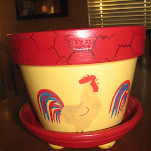 May include: A red and yellow ceramic planter with a rooster painted on the front. The rooster has a red comb and wattle, and its tail feathers are red, white, and blue. The planter has a red rim and base, and three small feet.