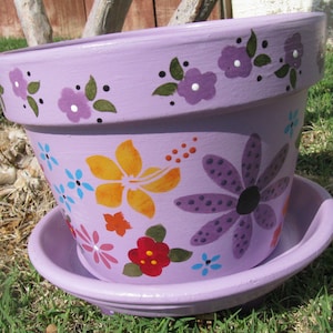 May include: A lavender-coloured terracotta flower pot with a saucer. The pot is decorated with painted flowers in shades of purple, yellow, orange, blue, and red.