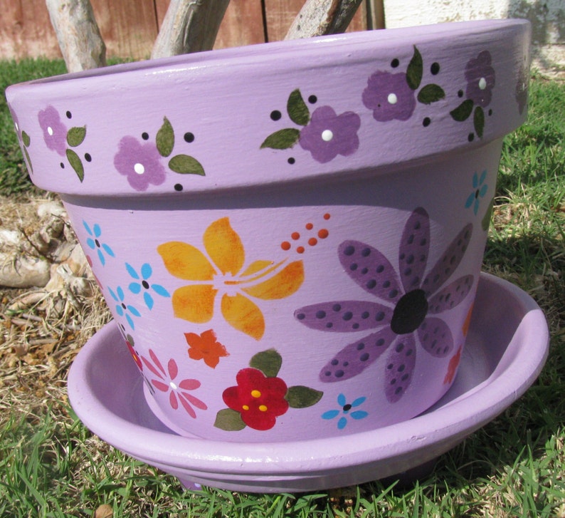 May include: A lavender-coloured terracotta flower pot with a saucer. The pot is decorated with painted flowers in shades of purple, yellow, orange, blue, and red.