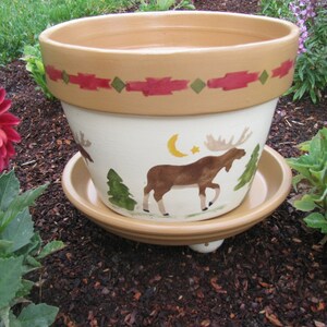 May include: A white ceramic planter with a brown rim and a moose design. The planter has a red and green geometric pattern around the top rim. The planter is sitting on a brown saucer.