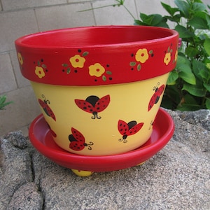 May include: A yellow and red terracotta flower pot with a ladybug design. The pot has a red rim and a saucer.