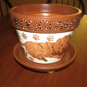May include: A brown and white ceramic planter with a dog illustration and the words "Doggy Days" written on the side. The planter has a matching brown saucer.