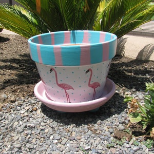 May include: A pink and blue striped terracotta planter with a white background and pink flamingo silhouettes. The planter has a saucer.