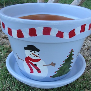 May include: A light blue ceramic planter with a white and red striped design around the top. The planter has a painted snowman and a Christmas tree on the front.