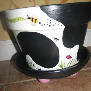 May include: Black and white ceramic planter with a cow print design. The planter has a saucer and pink feet. The planter is decorated with painted bees, ladybugs, and ants.