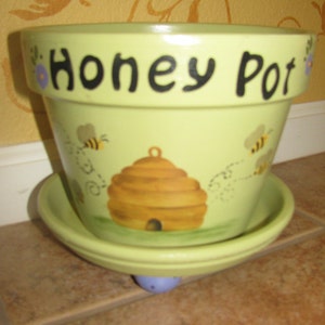 May include: A light green ceramic flower pot with a matching saucer. The pot has the words "HONEY POT" in black lettering. It features a painted beehive and bees. The pot is sitting on a tile surface.