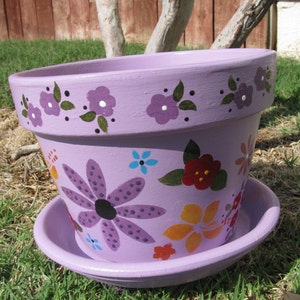 May include: A purple painted terracotta flower pot with a saucer. The pot is decorated with purple, red, yellow, and blue flowers. The pot has a white rim and a black rim.
