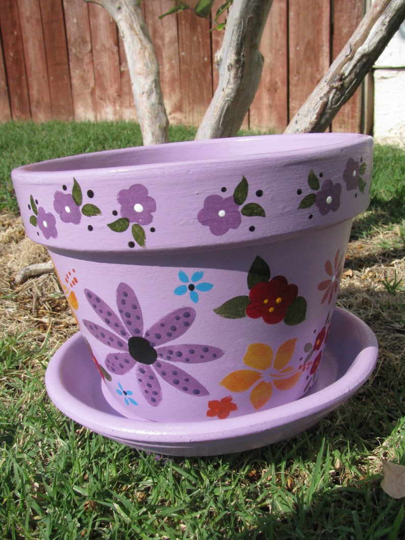 May include: A purple painted terracotta flower pot with a saucer. The pot is decorated with purple, red, yellow, and blue flowers. The pot has a white rim and a black rim.