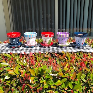 Könnte beinhalten: Fünf handbemalte Terrakotta-Blumentöpfe mit verschiedenen Designs. Die Töpfe sind in einer Reihe auf einer schwarz-weißen karierten Tischdecke angeordnet. Die Töpfe sind mit verschiedenen festlichen Motiven verziert, darunter Schneeflocken, Rentiere und Sterne. Die Töpfe stehen auf kleinen, dreibeinigen Ständern.