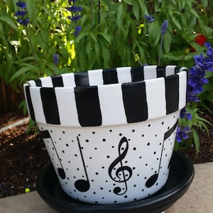 May include: A white ceramic planter with black stripes and musical notes painted on it. The planter has a black saucer with three feet.