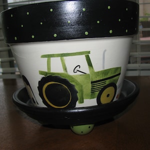 May include: A white ceramic planter with a black rim and green polka dots. The planter is decorated with a green tractor with black wheels and yellow rims. The planter has a black saucer with a green base.