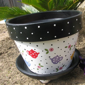 May include: A black and white polka dot planter with a white background and colorful teapots painted on it. The planter has a black saucer and a small foot.