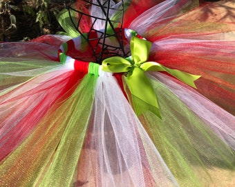 May include: A red, green, and white tulle tutu with a green satin bow. The tutu is made of multiple layers of tulle and has a white elastic waistband.