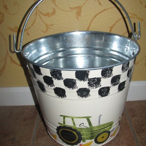 May include: A white metal bucket with a black and white checkered pattern. The bucket has a green tractor and yellow chicks painted on the side. The bucket has a silver handle.