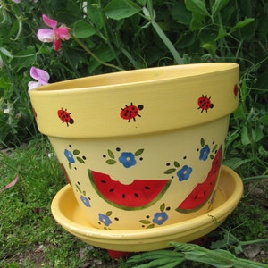 May include: Yellow ceramic planter with a painted design of watermelons, ladybugs, and blue flowers. The planter has a saucer.