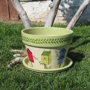 May include: A green and white ceramic planter with a leafy green trim and painted birdhouses. The birdhouses are painted in red, blue, and green.