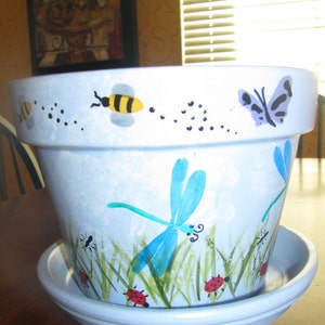May include: A light blue ceramic flower pot with a matching saucer. The pot is decorated with hand-painted insects, including bees, butterflies, dragonflies, ladybugs, and ants. The pot is approximately 7 inches in diameter.