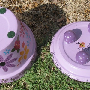 May include: Two purple painted terracotta flower pots. One pot has a floral design and three green felt pads on the bottom. The other pot has four purple polka dot feet and the word "Happy" painted on the bottom.