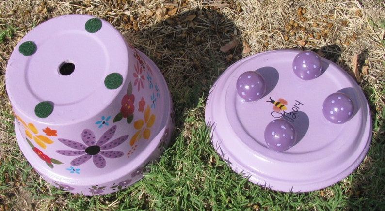 May include: Two purple painted terracotta flower pots. One pot has a floral design and three green felt pads on the bottom. The other pot has four purple polka dot feet and the word "Happy" painted on the bottom.
