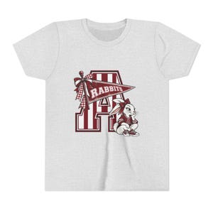 May include: A light heather gray t-shirt featuring a large maroon and white letter A with vertical stripes. A maroon pennant flag with the text RABBITS and a plaid bow is attached to the letter. A cartoon rabbit wearing a maroon shirt sits nearby.