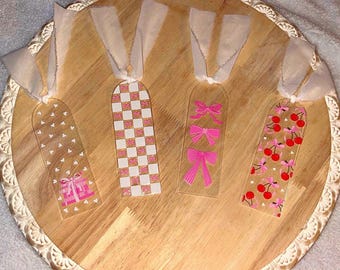 Può includere: Quattro segnalibri trasparenti in acrilico con nastri bianchi. I disegni includono fiocchi rosa, ciliegie, un regalo e un motivo a scacchi. I segnalibri sono disposti su un vassoio di legno.