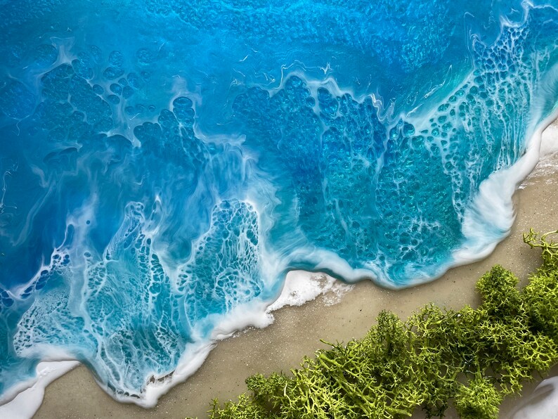 Pu&ograve; includere: Vista aerea di una scena di spiaggia con onde bianche e schiumose che si infrangono su una riva sabbiosa. L'oceano passa dal blu intenso al turchese, con muschio verde nell'angolo in basso a destra.