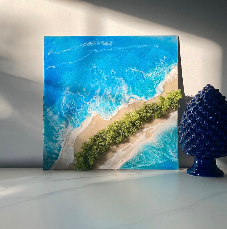 Pu&ograve; includere: Opera d'arte quadrata in resina raffigurante una scena di spiaggia. L'opera presenta una riva sabbiosa, vegetazione verde e onde blu dell'oceano. Il pezzo &egrave; esposto su una superficie bianca con un oggetto decorativo blu.