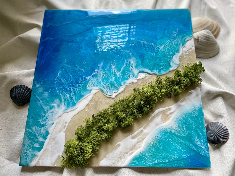 Pu&ograve; includere: Opera d'arte quadrata in resina raffigurante una scena di spiaggia. L'opera presenta vibranti onde blu e turchesi con schiuma bianca, una spiaggia sabbiosa e una striscia di muschio verde. Conchiglie sono posizionate attorno all'opera.